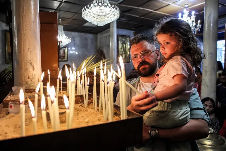 A Christian worshipper carrying a child lights a candle during the Palm Sunday service at the start of Holy Week for Orthodox Christians, at the Greek Orthodox Church of Saint Porphyrius in Gaza City on April 28, 2024, amid the continuing Israeli assault [Omar Al-Qattaa/AFP]